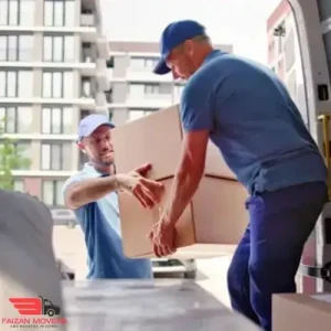 Movers and Packers in Meydan 10 Two workers of Faizan Movers and Packers in JVC, Dubai in blue uniforms are loading a large cardboard box into a van. They are outside an apartment building, collaborating efficiently.