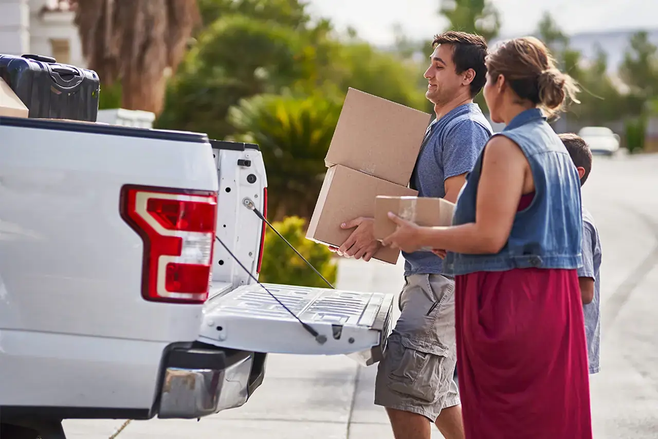 Best Pickup Truck for Rent in Dubai 7 A man and woman load cardboard boxes into a white 1 ton pickup truck rental on a sunny day. They appear to be moving, with greenery in the background.